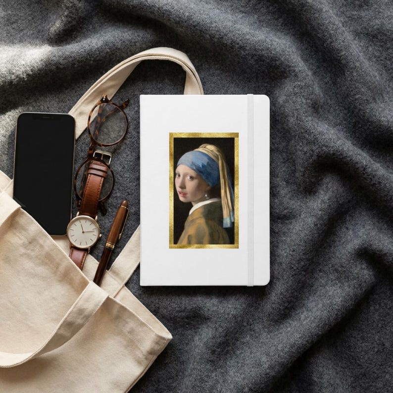 May include: A white notebook featuring a rectangular print of the Girl with a Pearl Earring painting framed in gold. The notebook is placed next to a black smartphone, a brown leather-strap wristwatch, and a brown pen inside a beige tote bag.