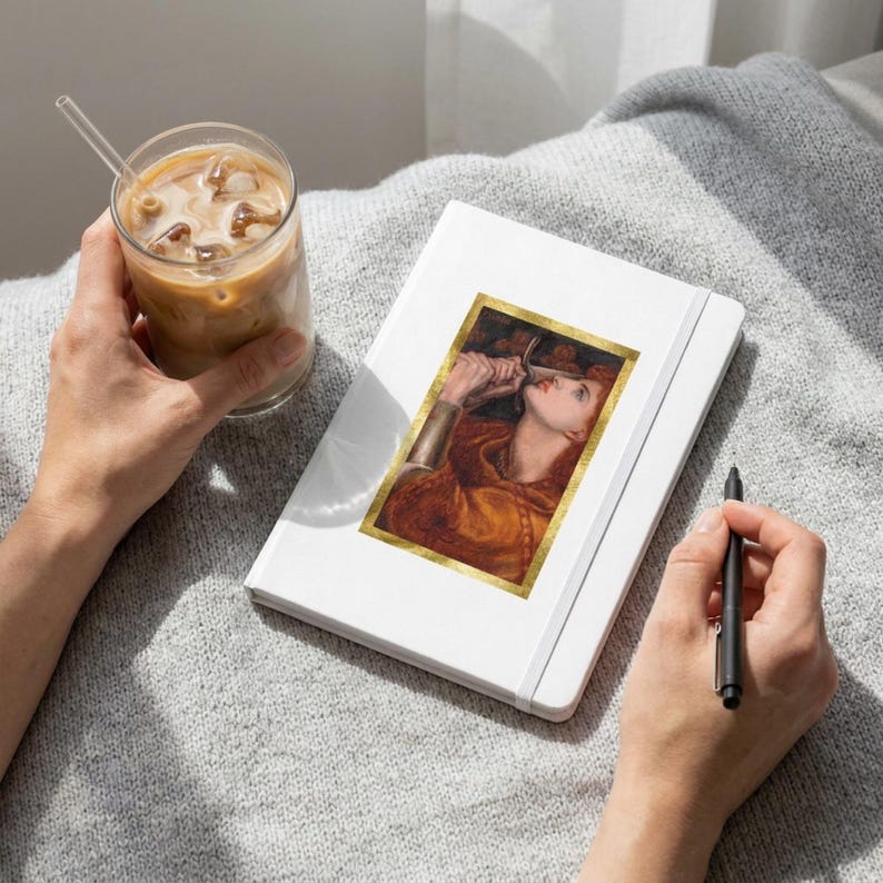 May include: A white notebook featuring a rectangular print of a painting depicting a person with long red hair holding a sword near their face, framed in gold. The notebook is held alongside a glass of iced coffee and a black pen.