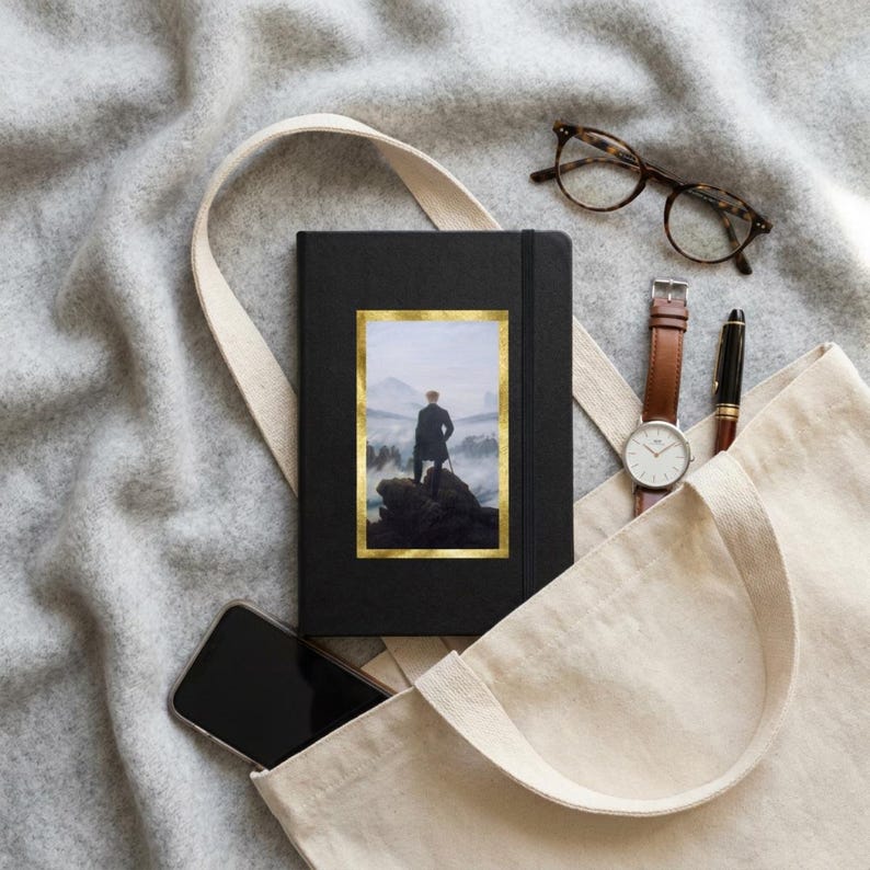 May include: A black notebook featuring a gold-framed print of a person standing on a rocky cliff overlooking a misty mountain landscape. The notebook is placed inside a beige canvas tote bag alongside a smartphone, a wristwatch, and eyeglasses.