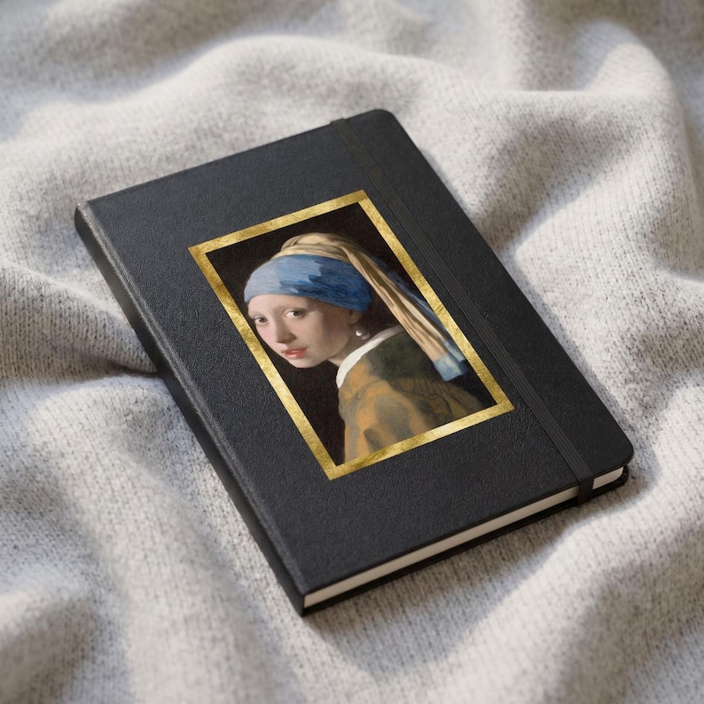 May include: A black hardcover notebook featuring a rectangular print of the painting Girl with a Pearl Earring, framed by a gold border. The notebook has a black elastic closure band and a textured cover, displaying the portrait of a person.