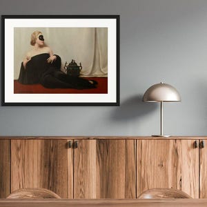 May include: A framed art print featuring a painting of a person with blonde hair wearing a black mask and a long black garment, reclining on a red surface against a white draped background, with a dark ornate vessel placed beside them.