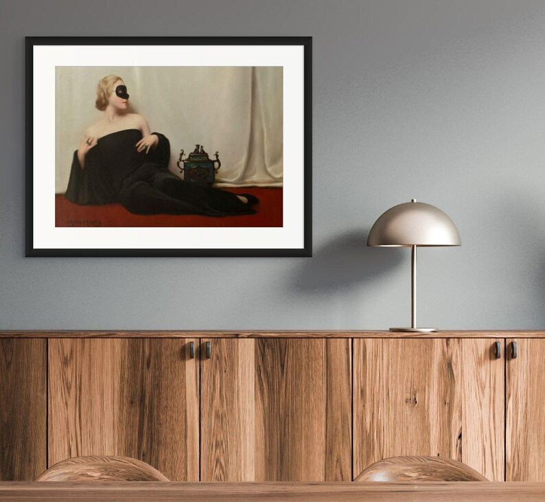 May include: A framed art print featuring a painting of a person with blonde hair wearing a black mask and a long black garment, reclining on a red surface against a white draped background, with a dark ornate vessel placed beside them.