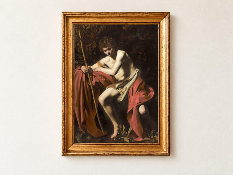 Saint John the Baptist in the Wilderness by Caravaggio, 1604, as a framed print in an antique gold frame on a cream wall. The young saint sits brooding in shadow, wrapped in a deep red cloak and holding a slender reed cross, lit by a single shaft of light.