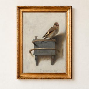 The Goldfinch by Carel Fabritius, 1654, as a framed print in an antique gold frame on a cream wall. A small goldfinch perches on its wooden feeder box, chained by the ankle, against a pale plaster wall signed C Fabritius 1654.