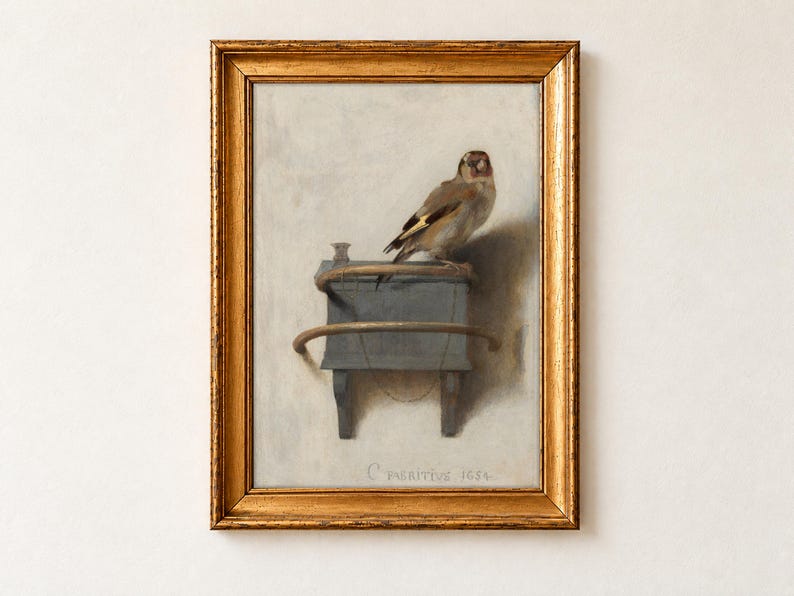 The Goldfinch by Carel Fabritius, 1654, as a framed print in an antique gold frame on a cream wall. A small goldfinch perches on its wooden feeder box, chained by the ankle, against a pale plaster wall signed C Fabritius 1654.