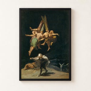 Witches' Flight by Francisco Goya, 1798, as a framed print in a black frame on a cream wall. Three witches in tall pointed hats fly through a black night sky carrying a man, while below one figure hides under a white sheet and another lies on the ground beside a donkey.