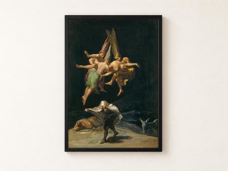 Witches' Flight by Francisco Goya, 1798, as a framed print in a black frame on a cream wall. Three witches in tall pointed hats fly through a black night sky carrying a man, while below one figure hides under a white sheet and another lies on the ground beside a donkey.