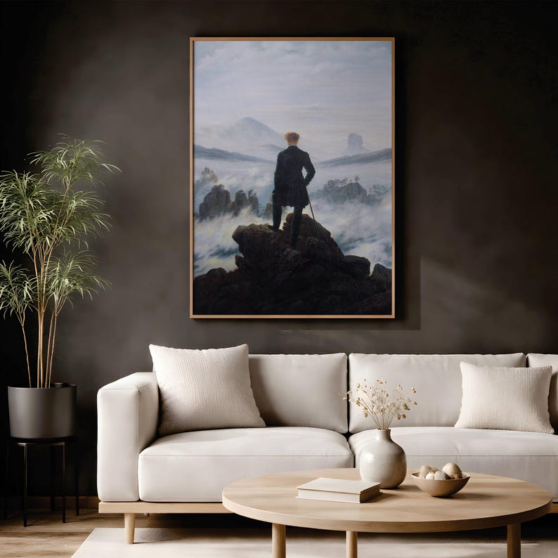 May include: A framed print of Caspar David Friedrich's Wanderer above the Sea of Fog, depicting a person in a dark coat standing on a rocky cliff overlooking a misty mountain landscape, displayed on a wall above a white sofa.