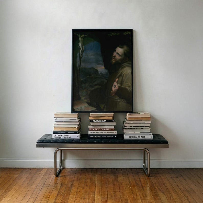 May include: A rectangular black-framed painting depicting a bearded figure in a brown robe looking toward a crucifix, mounted above a black tufted leather bench with a tubular metal frame, flanked by three stacks of various hardcover books.