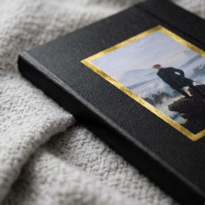 May include: A black hardcover book featuring a rectangular inset print of a person standing on a rocky cliff overlooking a misty landscape. The print is framed by a thin, reflective gold border that contrasts against the textured black cover material.