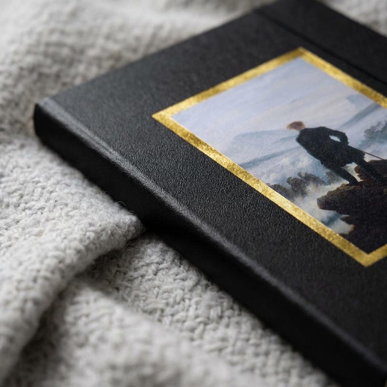 May include: A black hardcover book featuring a rectangular inset print of a person standing on a rocky cliff overlooking a misty landscape. The print is framed by a thin, reflective gold border that contrasts against the textured black cover material.