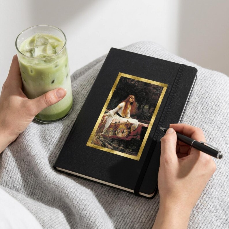 May include: A black hardcover notebook featuring a rectangular print of John William Waterhouse's painting The Lady of Shalott on the front cover, held by a person with a glass of iced matcha latte and a black ballpoint pen in their hands.