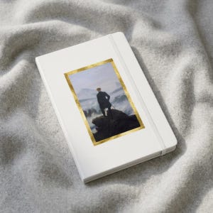 May include: A white notebook with an elastic closure band featuring a rectangular print of a person standing on a rocky cliff overlooking a misty mountain landscape, framed by a textured gold border on the front cover of the notebook.