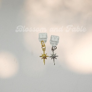May include: A pair of earrings with a starburst design. One earring is gold-colored, and the other is silver-colored. The earrings are hanging from clear plastic earring backs. The text "Blossom and Fable" is in the background.