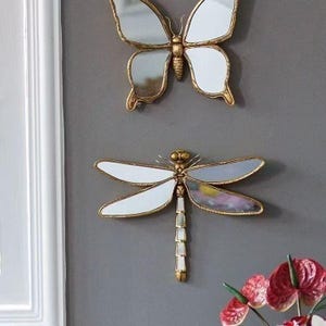 May include: Two decorative wall mirrors: a butterfly and a dragonfly. The butterfly mirror has a gold frame and white mirrored wings. The dragonfly mirror also has a gold frame with white and mirrored wings. The mirrors are mounted on a gray wall.