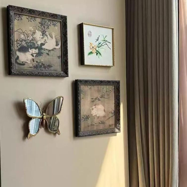 May include: A collection of framed artwork and a butterfly-shaped mirror adorn a wall. The artwork includes a painting of cats, a floral design, and a cat. The butterfly mirror has a gold frame.