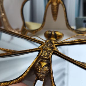 May include: Close-up of a gold-colored dragonfly-shaped mirror with intricate detailing. The dragonfly's wings are outstretched, and the body is textured. The mirror reflects the surrounding environment. The overall design is ornate and decorative.