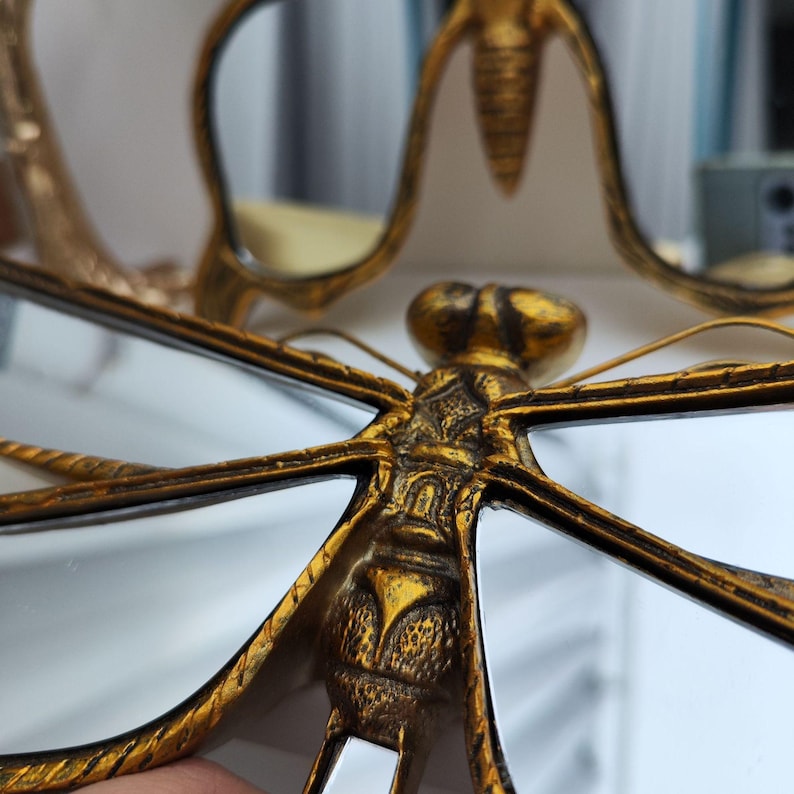 May include: Close-up of a gold-colored dragonfly-shaped mirror with intricate detailing. The dragonfly's wings are outstretched, and the body is textured. The mirror reflects the surrounding environment. The overall design is ornate and decorative.