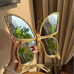 Puede incluir: Espejo decorativo con forma de mariposa y marco dorado. El espejo tiene cuatro alas espejadas y un cuerpo detallado. El marco presenta un aspecto texturizado y tallado. Un objeto decorativo.