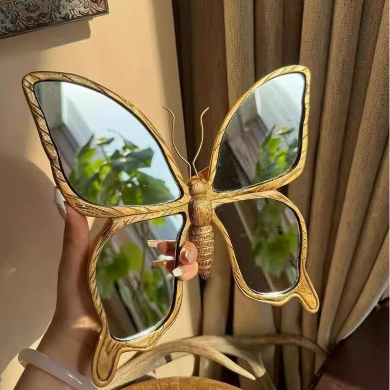 May include: A decorative butterfly mirror with a gold-colored frame. The mirror is shaped like a butterfly with four mirrored wings and a detailed body. The frame has a textured, carved appearance, and the mirror reflects the surrounding environment.