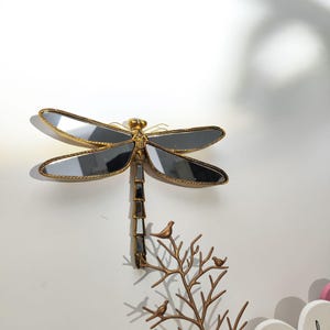 May include: A decorative dragonfly wall hanging with mirrored wings and a gold-colored frame. The dragonfly is mounted above a bronze-colored tree branch with three small bird figurines. The wall is white.