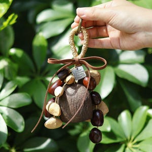 May include: A handmade charm featuring a cluster of natural seeds and nuts in shades of brown and beige, suspended from a woven loop. A small metal tag with the text "Follow your dreams, trust your heart" is attached with a brown leather cord.