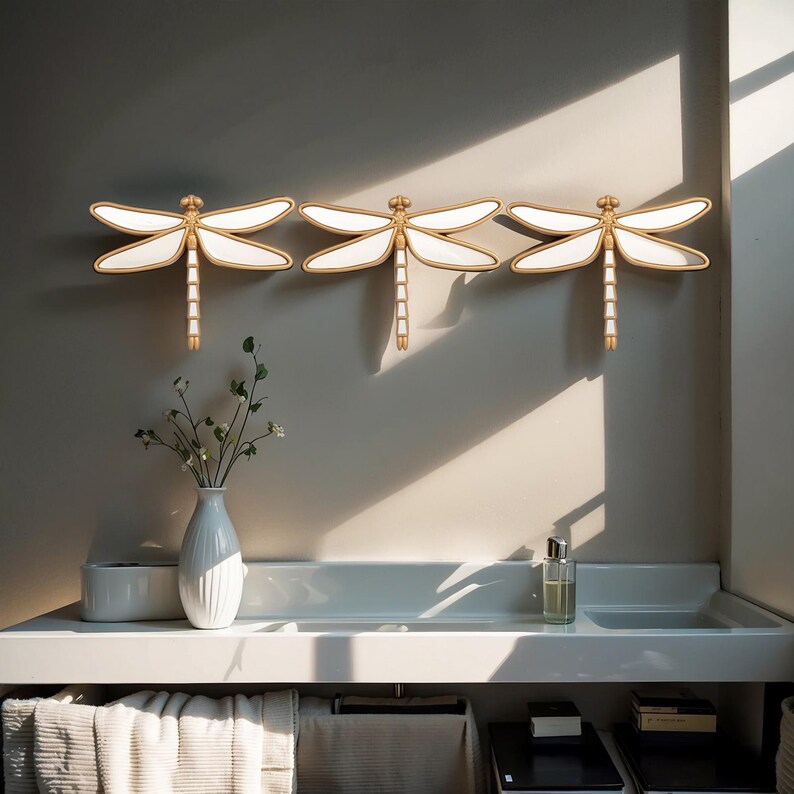 May include: Three decorative dragonfly wall sconces. Each dragonfly features white wings with gold trim and a gold body. The sconces are mounted on a neutral-colored wall above a white countertop with a vase of flowers and other bathroom accessories.