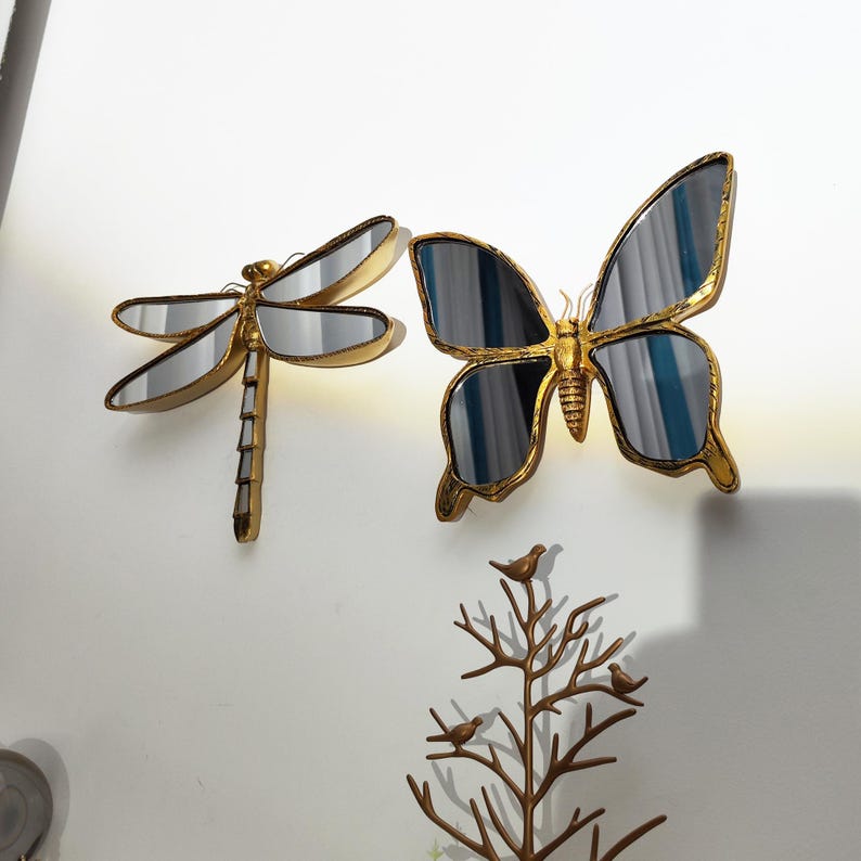 May include: Gold-colored metal dragonfly and butterfly wall mirrors with mirrored surfaces. The dragonfly has a segmented body and wings, while the butterfly has large, detailed wings. A decorative metal tree with birds is also present.