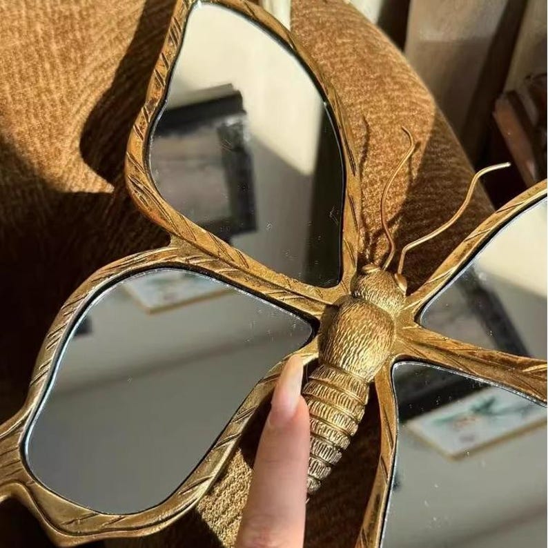 May include: A gold-colored butterfly-shaped mirror with multiple reflective surfaces. The mirror has a detailed, textured frame and antenna-like elements. The butterfly's body is also intricately designed.