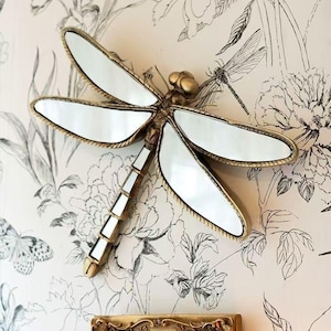 May include: A decorative dragonfly mirror with a gold-colored frame and mirrored wings. The dragonfly is mounted on a wall with a floral wallpaper background. The mirror adds a touch of nature-inspired decor.