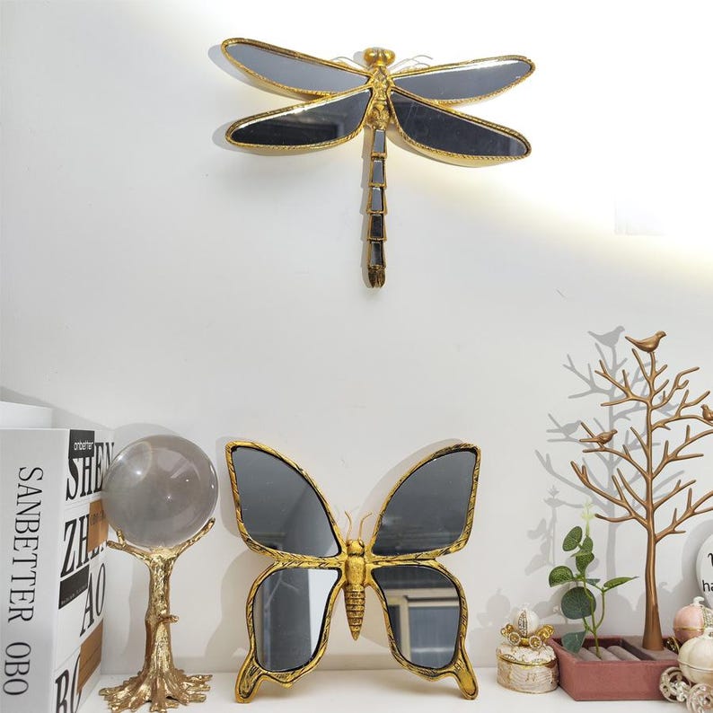 May include: A gold-framed dragonfly mirror and a butterfly mirror with dark gray wings are mounted on a white wall. A gold-colored decorative tree and a crystal ball are on a shelf below.