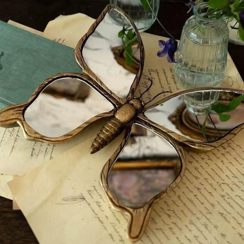 May include: A decorative butterfly-shaped mirror with a gold-colored frame and reflective surfaces. The mirror is resting on a vintage paper surface, with small glass vases and flowers in the background. The butterfly's body is detailed with textured lines.