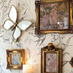 May include: A wall display featuring a butterfly-shaped mirror and several framed pictures with ornate gold frames. The pictures depict scenes with people. The wallpaper has a floral design in black and white.