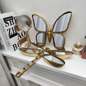 Puede incluir: Una colección de objetos decorativos, que incluye un espejo de mariposa dorado, un espejo de libélula y una bola de cristal sobre un soporte dorado. Los espejos de mariposa y libélula tienen superficies reflectantes. Los artículos se exhiben sobre una superficie blanca.