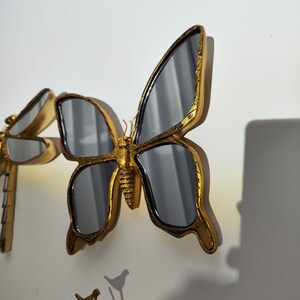 May include: A golden butterfly mirror with a detailed frame and reflective surface. The butterfly is mounted on a wall, showcasing its intricate design and elegant form. The mirror adds a touch of nature-inspired decor.
