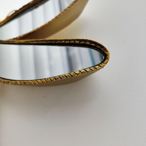 May include: Close-up of a decorative gold-colored mirror with a textured border. The mirror has an oval shape and reflects a blurred image. The background is a plain white surface.