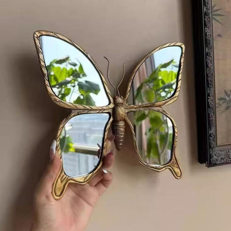 May include: A decorative butterfly mirror with a gold-colored frame. The mirror is shaped like a butterfly with four reflective surfaces. The frame has an ornate, textured design. The mirror is being held up by a hand.