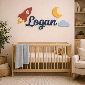 May include: A wooden crib with a light blue blanket and a teddy bear inside. Above the crib, wall decor features the name Logan in dark blue script, a red rocket ship with yellow flames, a yellow crescent moon, and a light blue cloud.