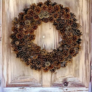 May include: A brown pine cone wreath hangs on a wooden door with red trim. The wreath is made of natural pine cones and is a festive decoration for the holiday season.