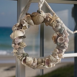 May include: A seashell wreath made of various sizes and shapes of seashells, starfish, and coral. The wreath is hanging on a window with a view of the ocean.