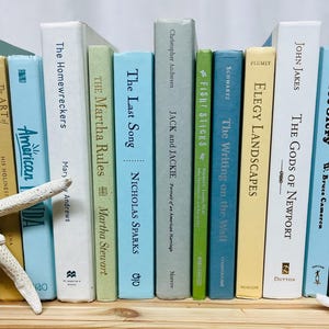 May include: A bookshelf with a variety of books stacked on top of each other. The books are all different colors and have different titles. There are two seashells on the bottom shelf.