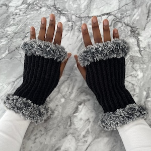 May include: A pair of fingerless gloves featuring a black ribbed knit texture. Each glove is accented with a thick, fluffy faux fur trim in shades of gray and white at both the wrist and the knuckles, providing a textured contrast to the knit.