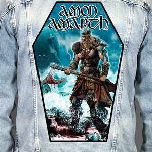 Può includere: Una giacca di jeans azzurra con una grande toppa posteriore a forma di bara. La toppa raffigura un guerriero vichingo che impugna un'ascia insanguinata, con la scritta AMON AMARTH in alto. La scena include un mare in tempesta, fulmini e una nave vichinga.