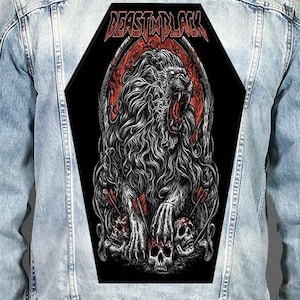 Può includere: Una giacca di jeans azzurra con una grande toppa posteriore a forma di bara. La toppa mostra un'illustrazione dettagliata di un leone ruggente con criniera fluente, teschi e frecce, con il testo BEAST IN BLACK stampato in rosso in alto.