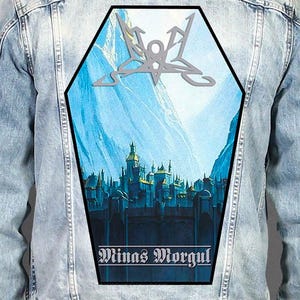 May include: A light blue acid-wash denim jacket featuring a large, coffin-shaped back patch. The patch displays a blue-toned illustration of a fortress with the text 'Minas Morgul' at the bottom and a silver, angular symbol at the top.