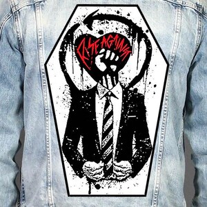 May include: A light blue denim jacket featuring a large, coffin-shaped graphic on the back. The print displays a black and white illustration of a person in a suit with a fist for a head, surrounded by a circular arrow and the red text Rise Against.