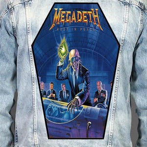 Peut inclure: Une veste en jean bleu clair avec un grand écusson dorsal en forme de cercueil représentant la pochette de l'album Rust in Peace de Megadeth. L'illustration montre un squelette en costume tenant un objet vert brillant au-dessus d'une capsule.