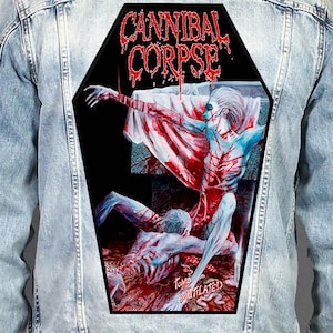 Peut inclure: Une veste en jean bleu clair avec un grand écusson dorsal en forme de cercueil. L'écusson affiche le texte CANNIBAL CORPSE en lettres rouges dégoulinantes au-dessus d'une illustration graphique de deux personnages dans une scène sanglante.