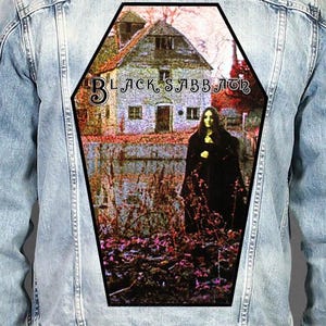 May include: A light blue denim jacket featuring a large, coffin-shaped back patch. The patch displays the Black Sabbath album cover art, showing a stone building and a figure in a black cloak standing in a field with purple and brown foliage.