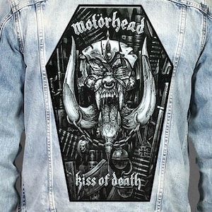 May include: A light blue denim jacket featuring a large, coffin-shaped back patch. The patch displays a black and white illustration of the Motörhead Snaggletooth mascot, with the text "motörhead" at the top and "kiss of death" at the bottom.